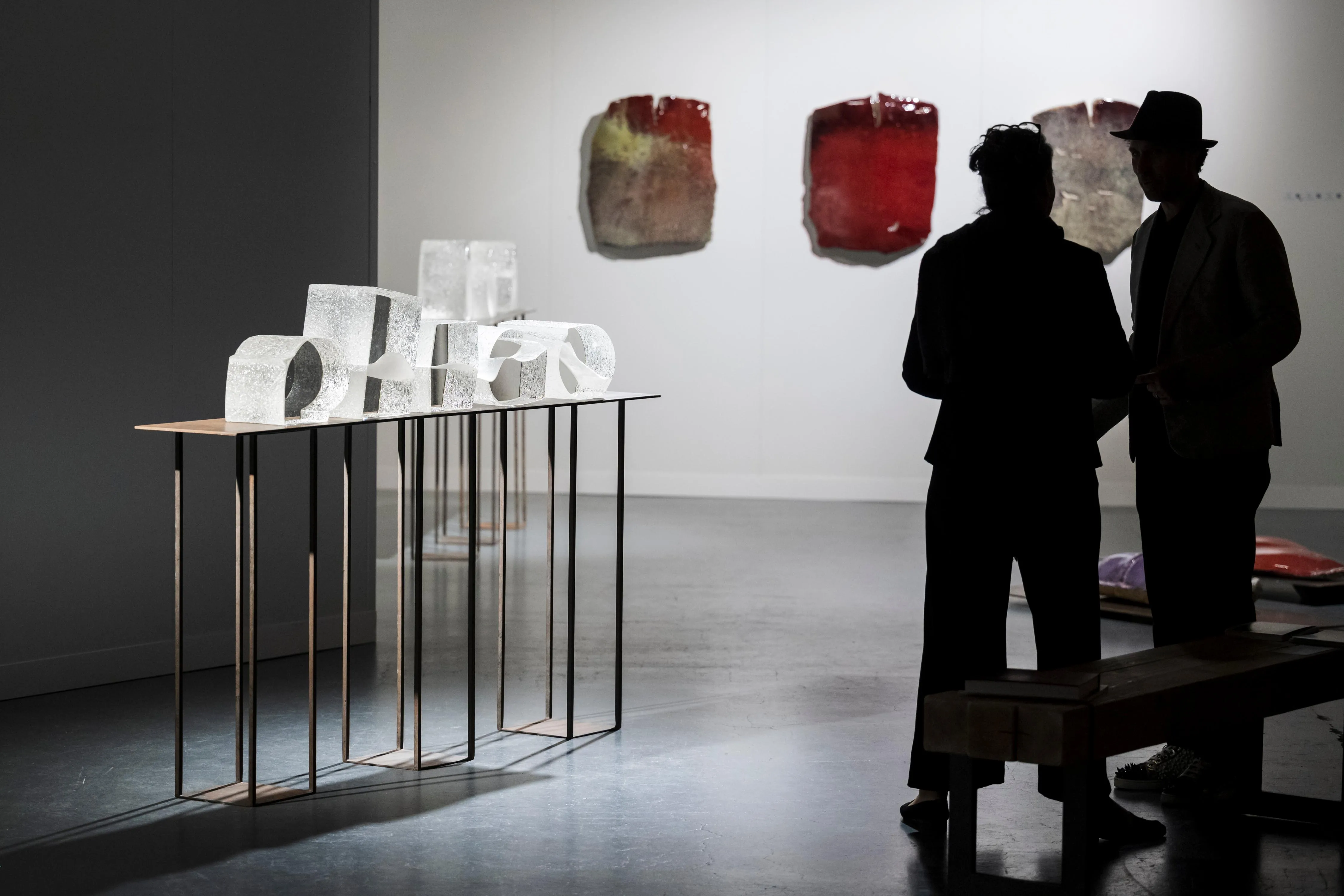 Translucent glass sculptures lined up on slender metal consoles, two visitors in silhouette in front of red wall pieces.