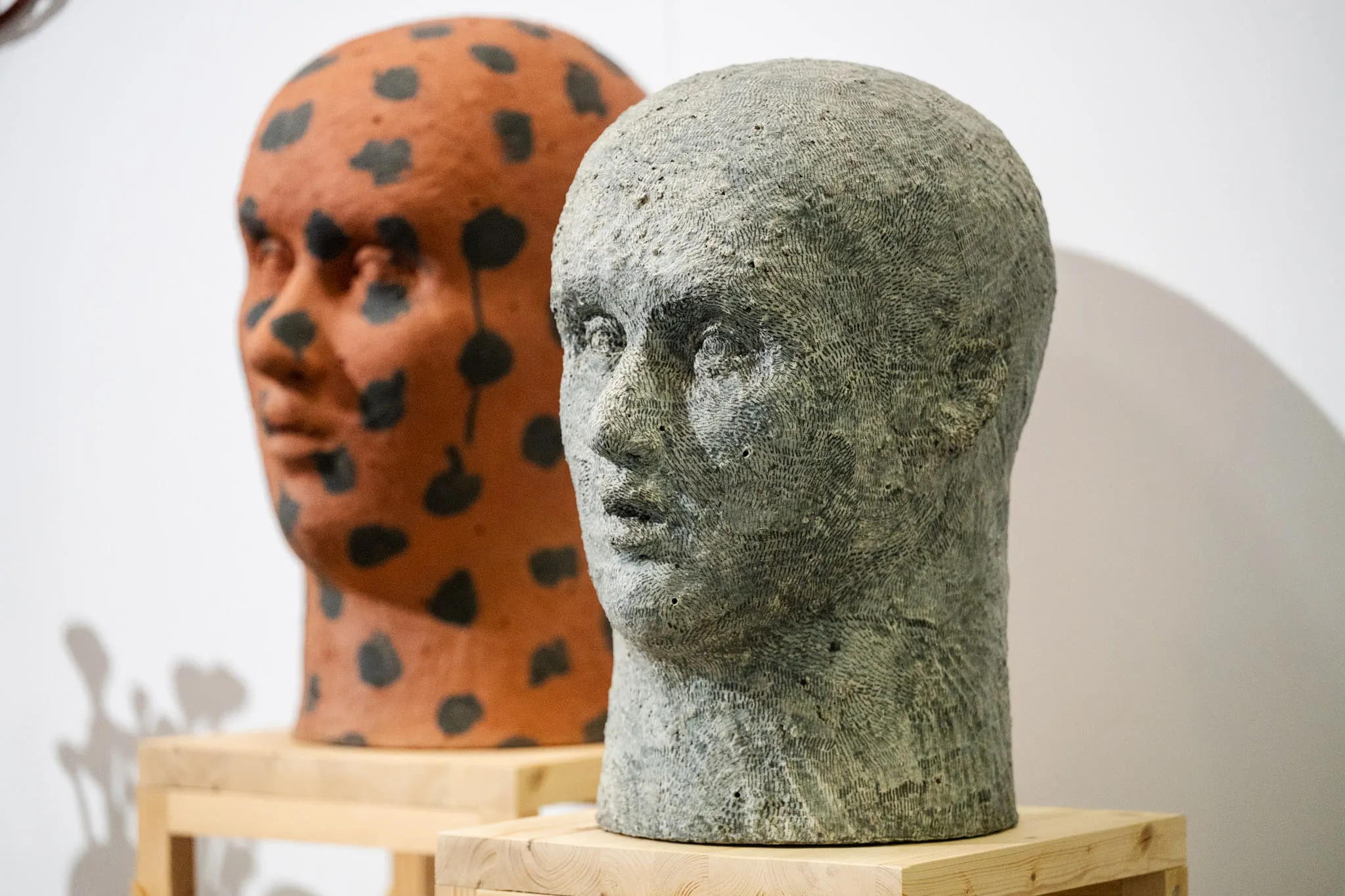 Two ceramic heads on wooden plinths, one grey, the other in terracotta flecked with black.