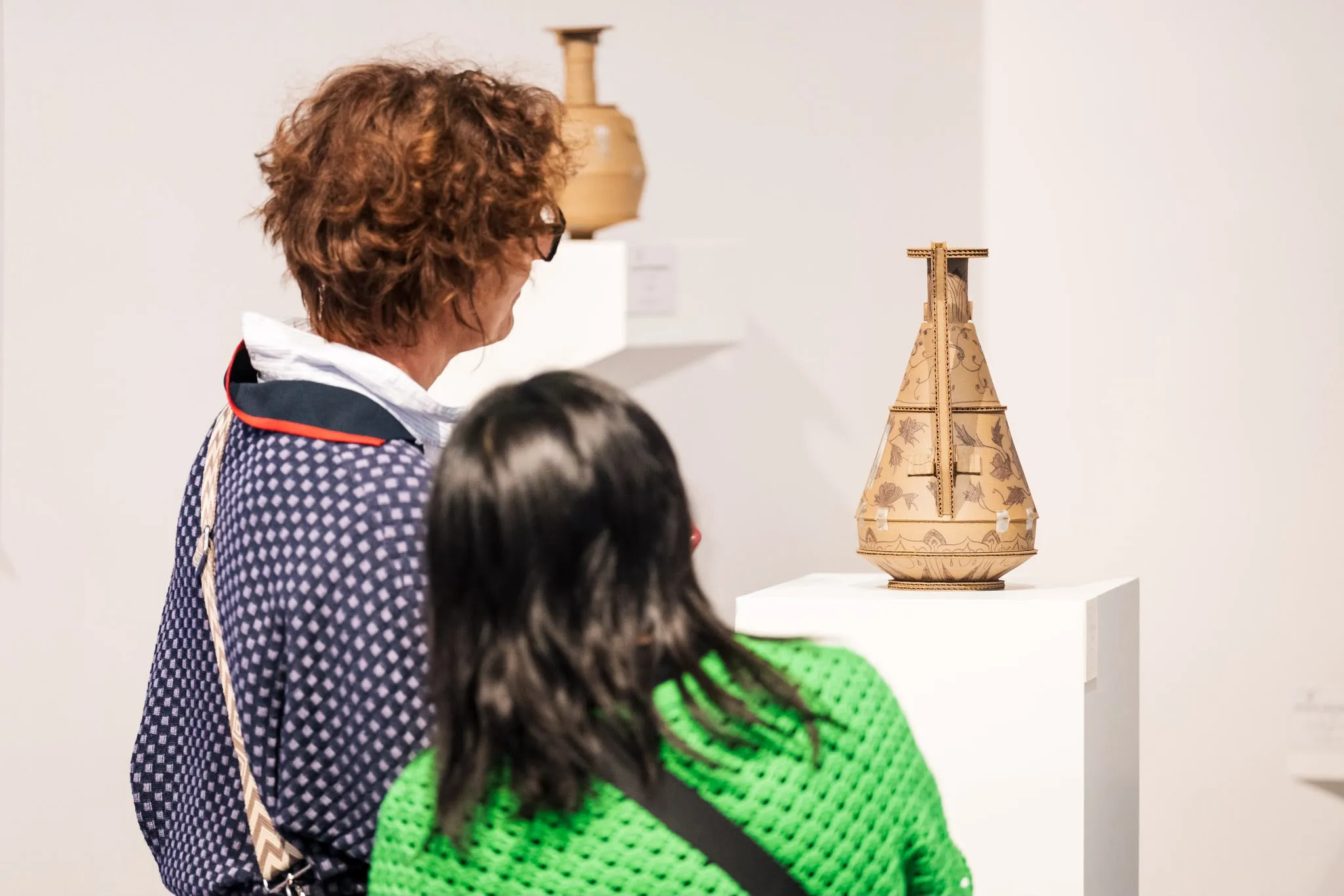 Two visitors looking at a cardboard vase with drawn decoration, set on a white plinth.