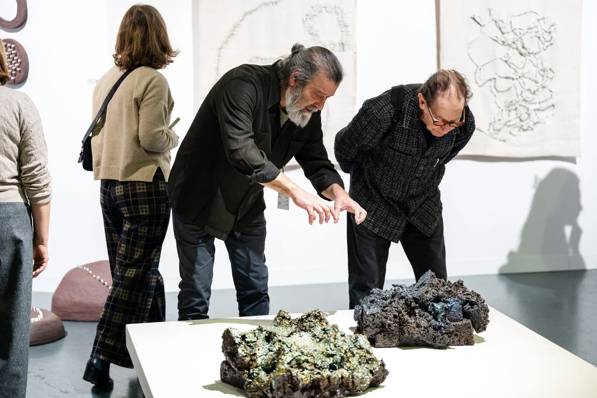 Two visitors leaning over a pair of ceramic sculptures on a white plinth.
